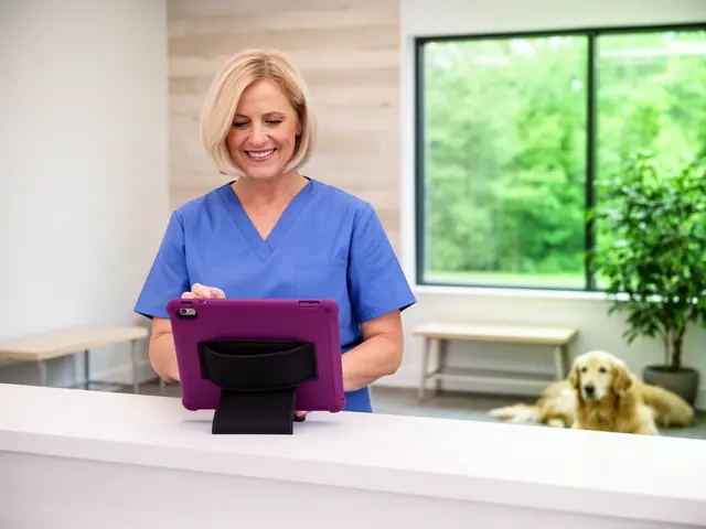 Smiling woman in blue scrubs using a plum tablet at a white reception counter, a golden retriever resting behind